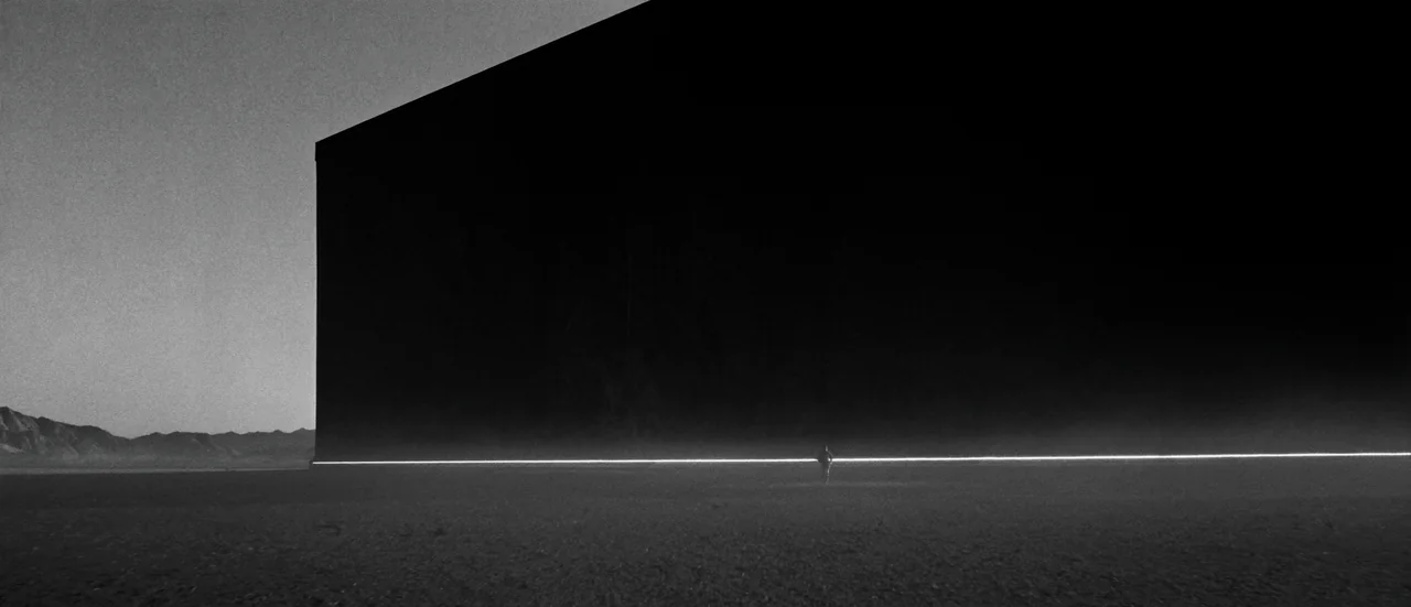 A vast dark building on a desert floor, a line of light along its base and a lone figure in front of it.