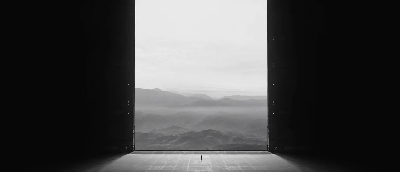 A lone figure stands at the threshold of a vast open door, with mountains and sky beyond.