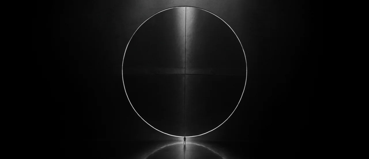 A circular door outlined in light at the end of a dark hall, with a lone figure at its threshold.