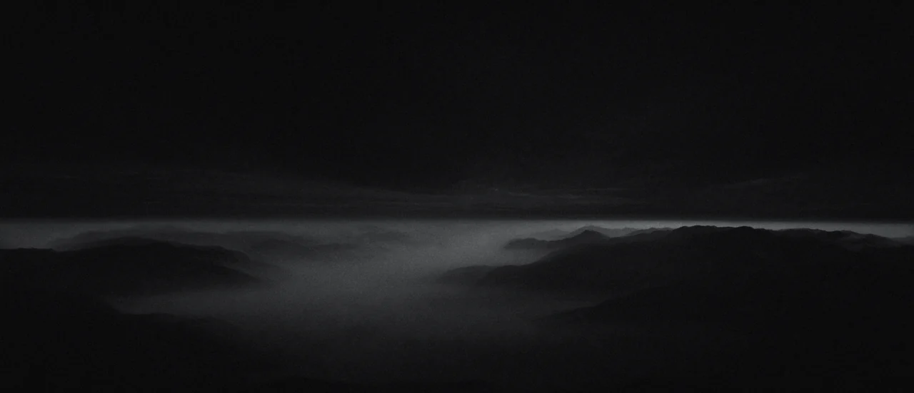 Mountain ridgelines under low fog at night, lit faintly along the horizon.
