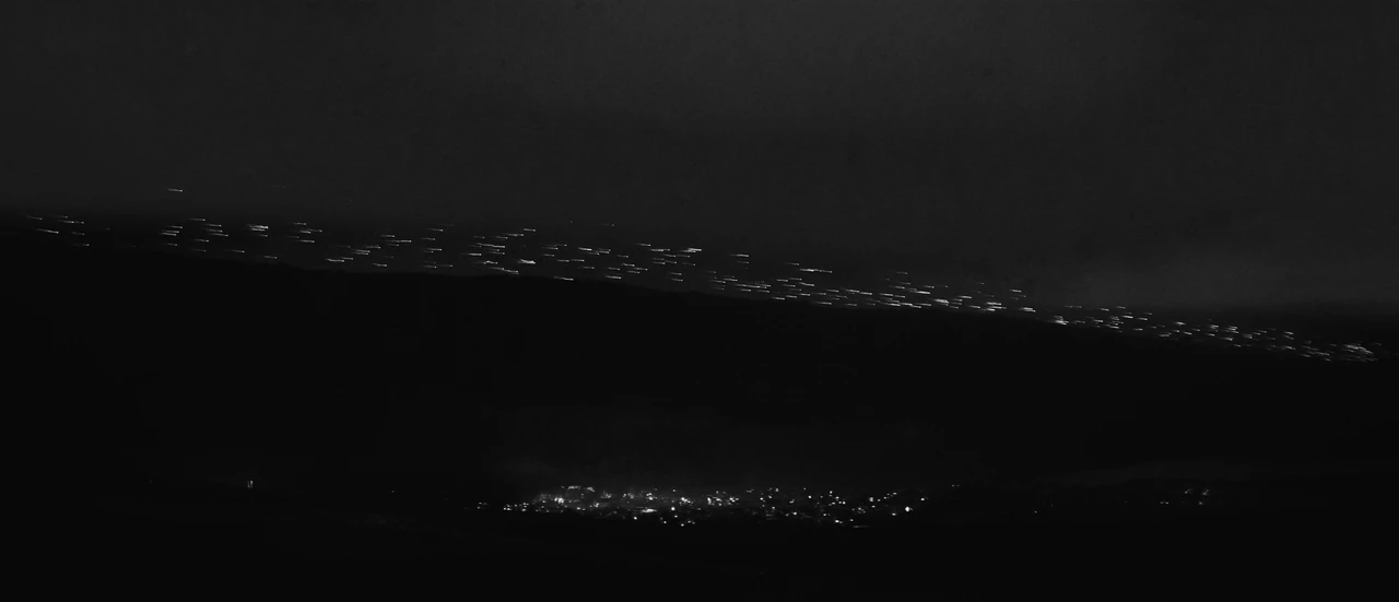 Many small lights cross the night sky low over a far ridge toward a sleeping town.