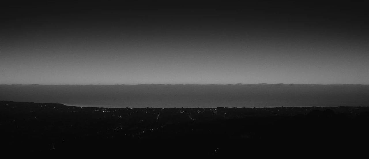 A coastal city before dawn, seen from a distant hill, with a bank of fog lying offshore.
