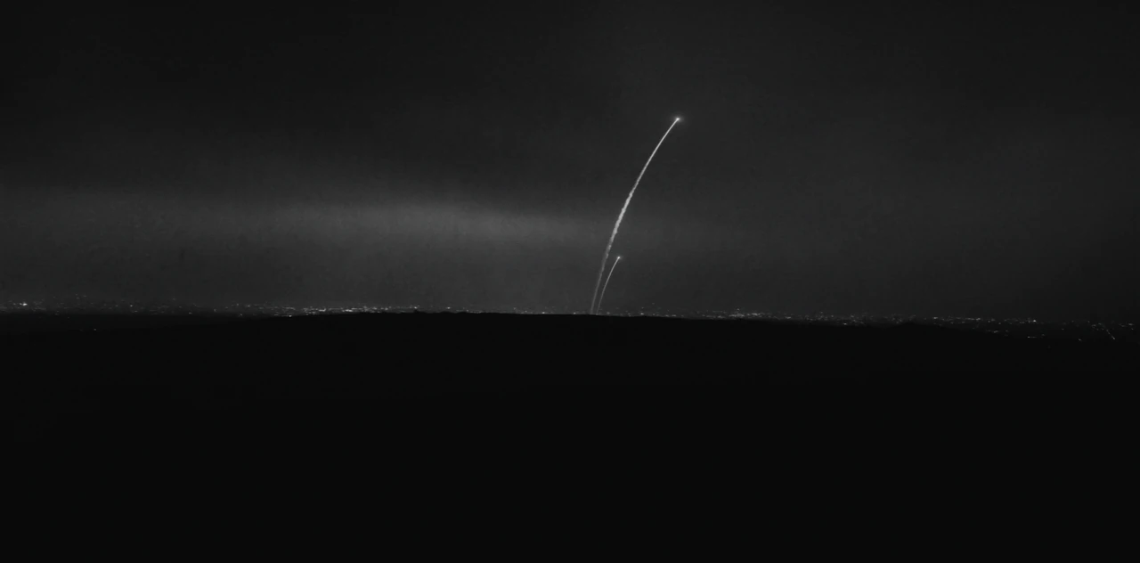 Thin trails of light rise from behind a dark ridge into the night sky, with faint lights along the horizon.