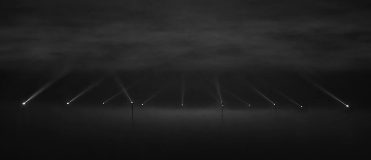 A row of small lights on thin masts across a fog-covered plain at night, each casting a faint beam up into a low cloud.