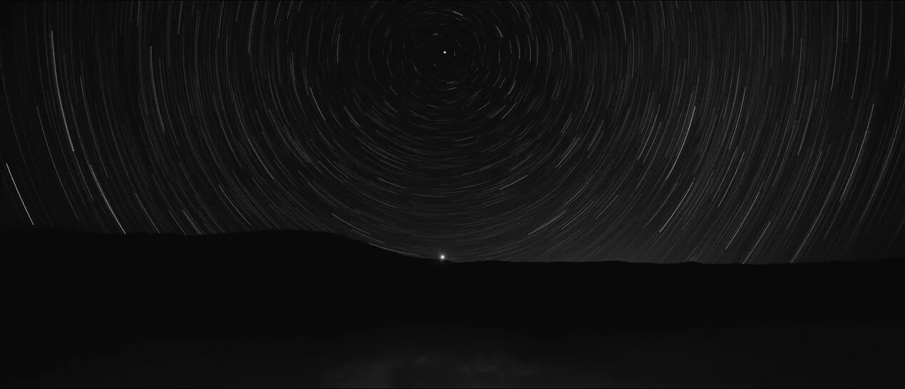 Stars trace circles around the pole star above a dark ridge with one small steady light on its crest.